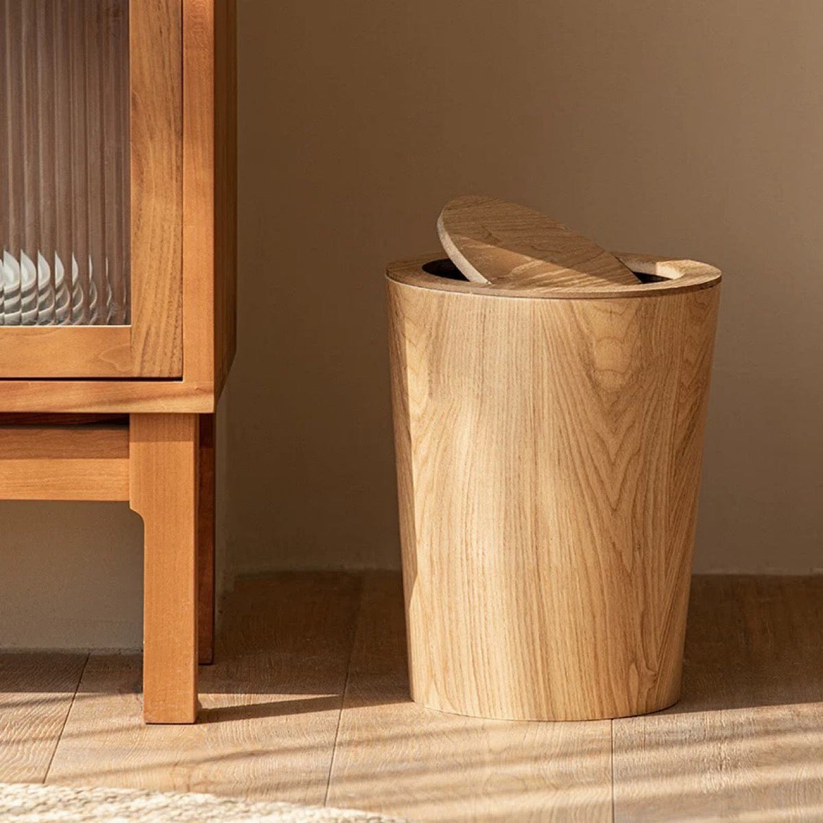 Natural wood grain trash can beside a wooden glass-front cabinet